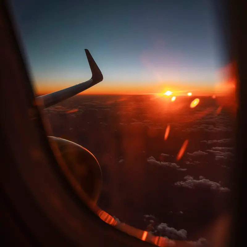 View through an aeroplane window at sunset, a dark wingtip against an orange horizon above the clouds