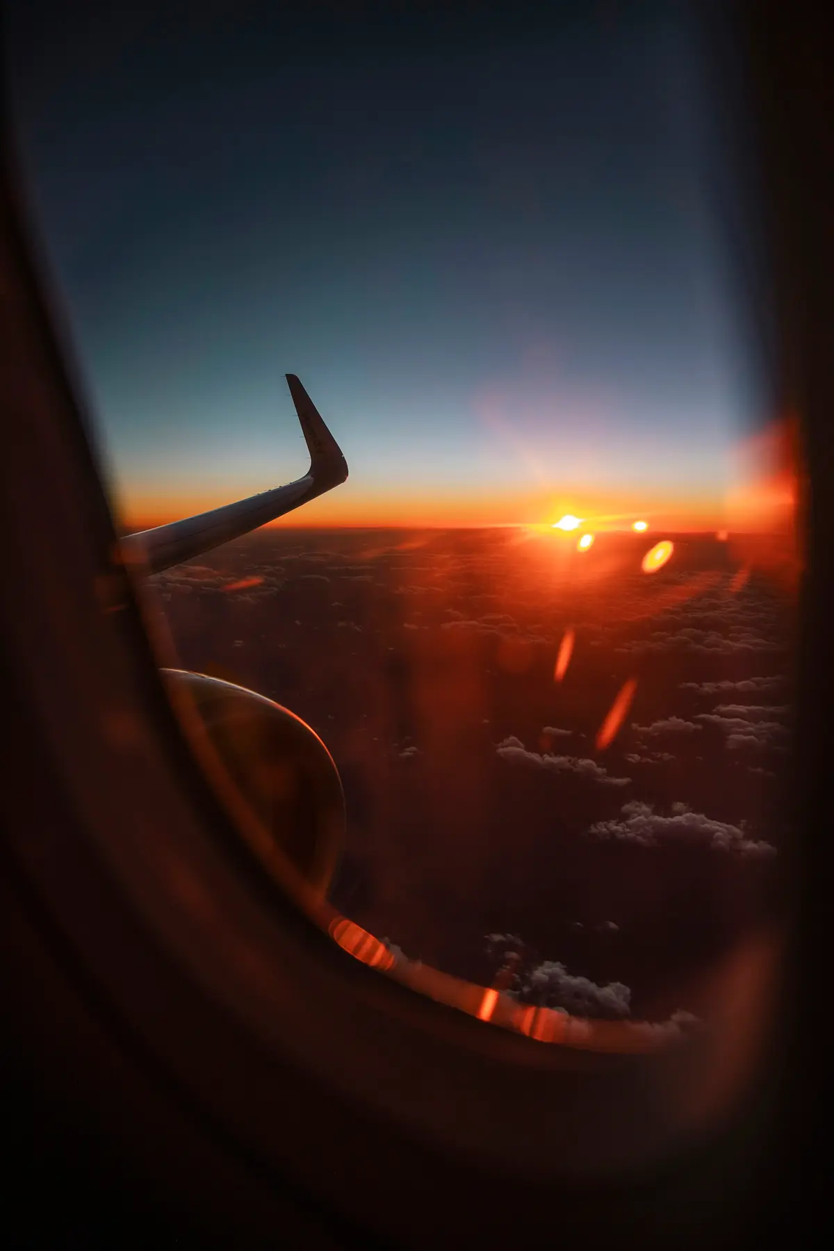 View through an aeroplane window at sunset, a dark wingtip against an orange horizon above the clouds