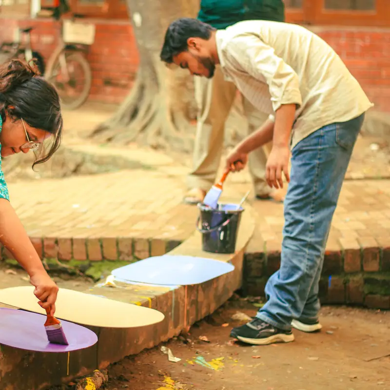 Two art students at Dhaka University painting large cut-out shapes in purple and cream on a brick ledge under trees