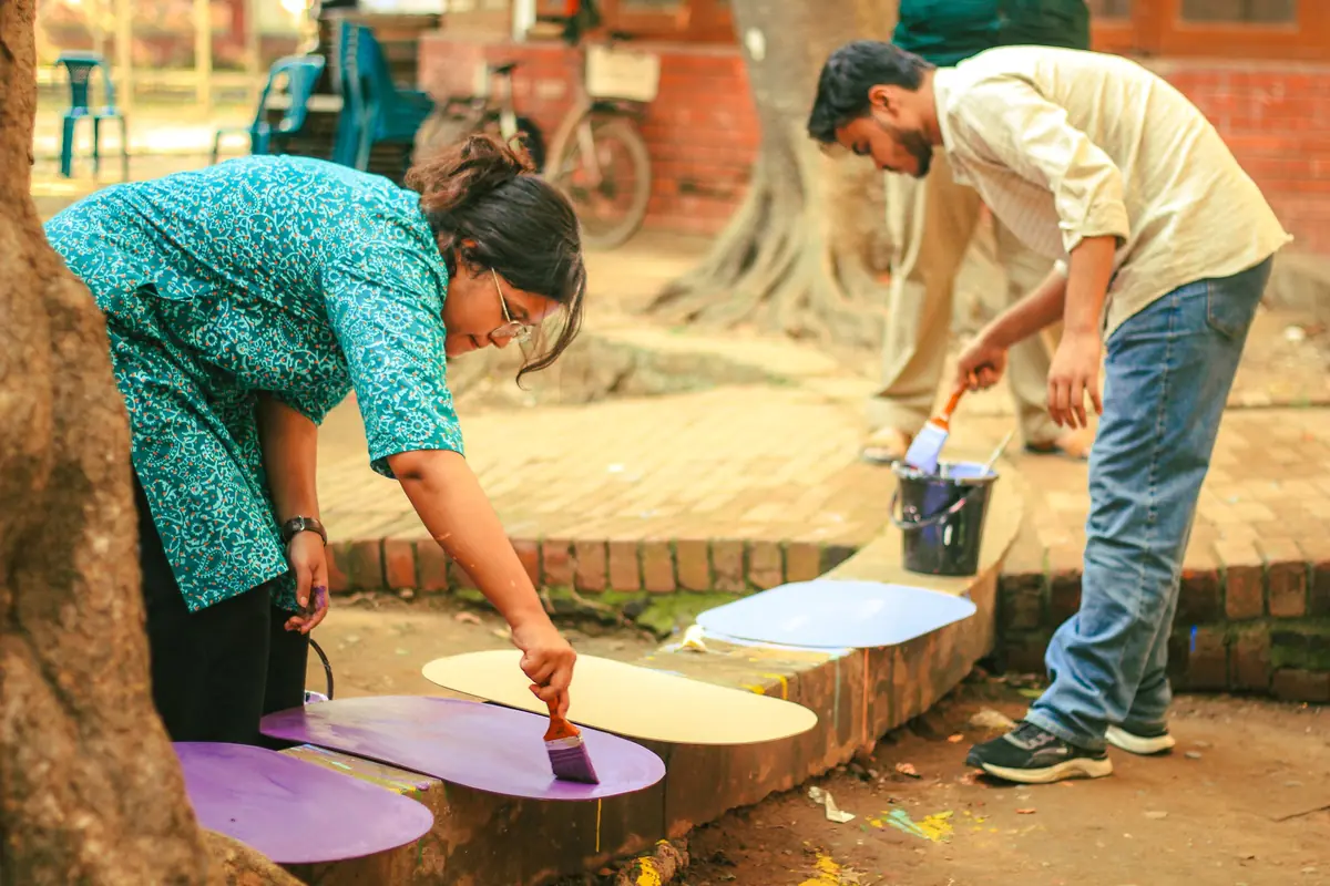 Two art students at Dhaka University painting large cut-out shapes in purple and cream on a brick ledge under trees