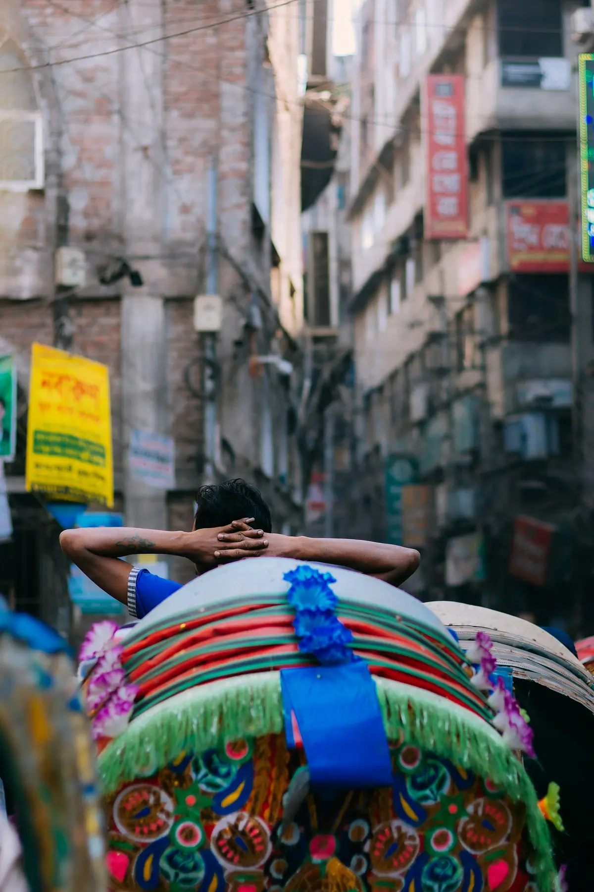 A rickshaw driver seen from behind, resting with his hands behind his head above a painted, flower-trimmed rickshaw hood in a narrow Dhaka street