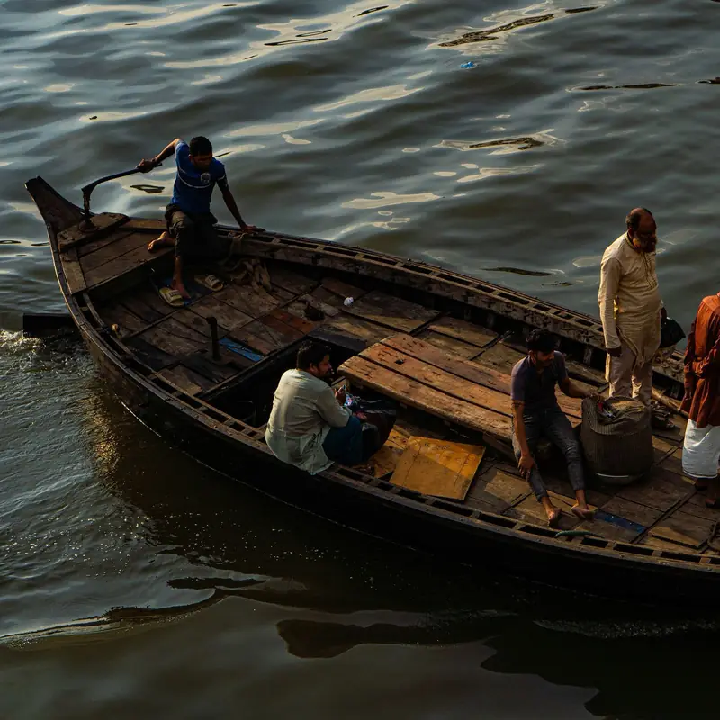 A long wooden boat carrying six men across dark, rippling water on the Buriganga River in Dhaka, in low golden light