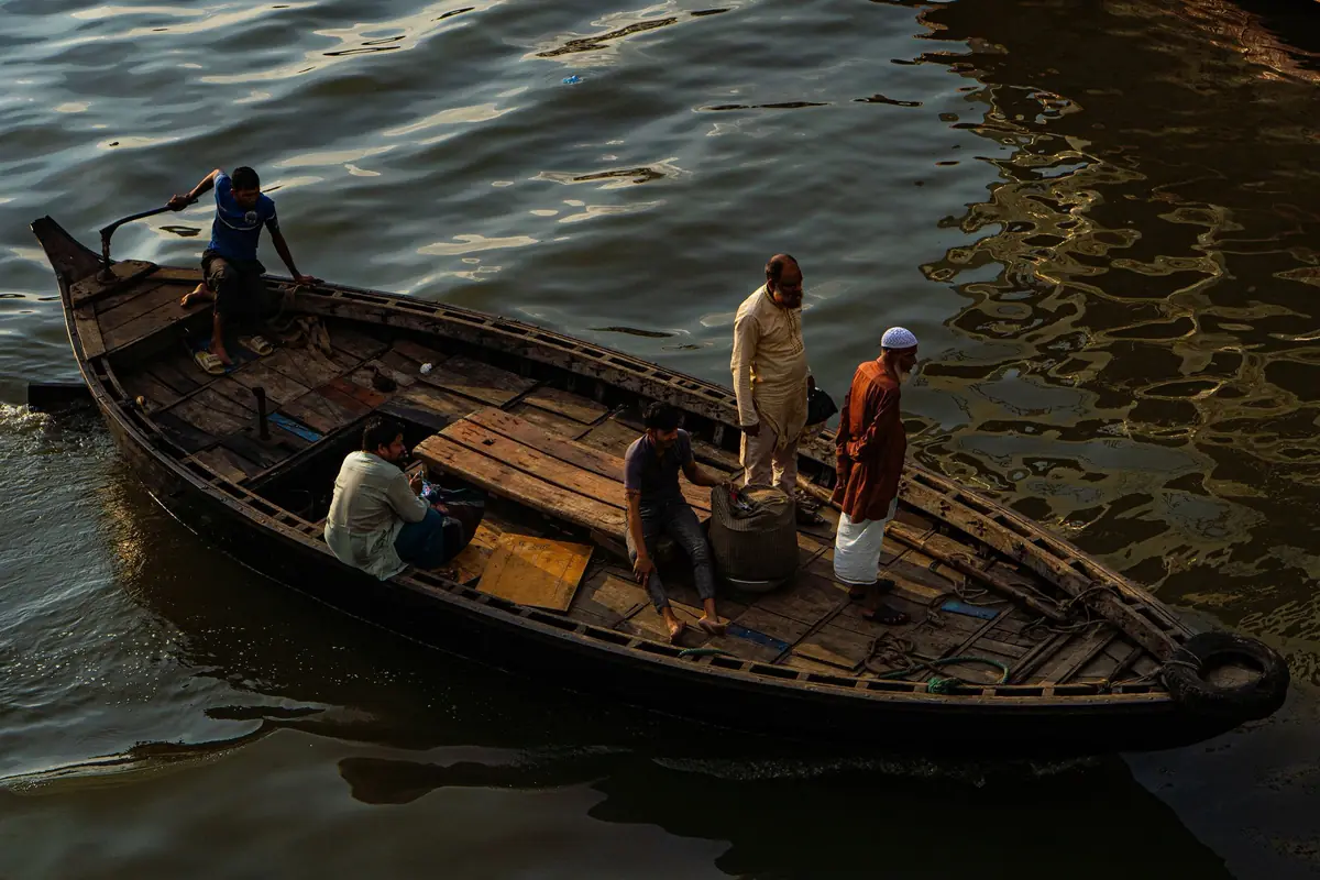 A long wooden boat carrying six men across dark, rippling water on the Buriganga River in Dhaka, in low golden light
