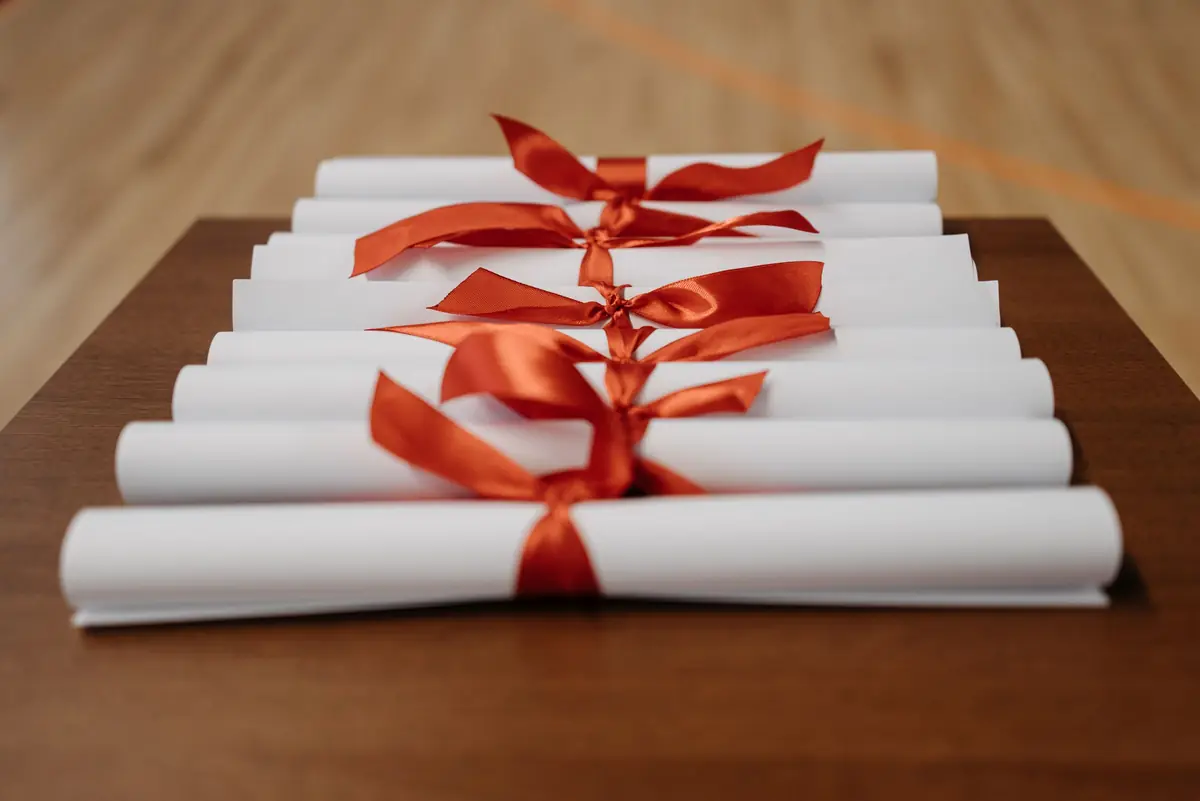 A row of rolled white diplomas, each tied with an orange-red satin bow, on a wooden desk