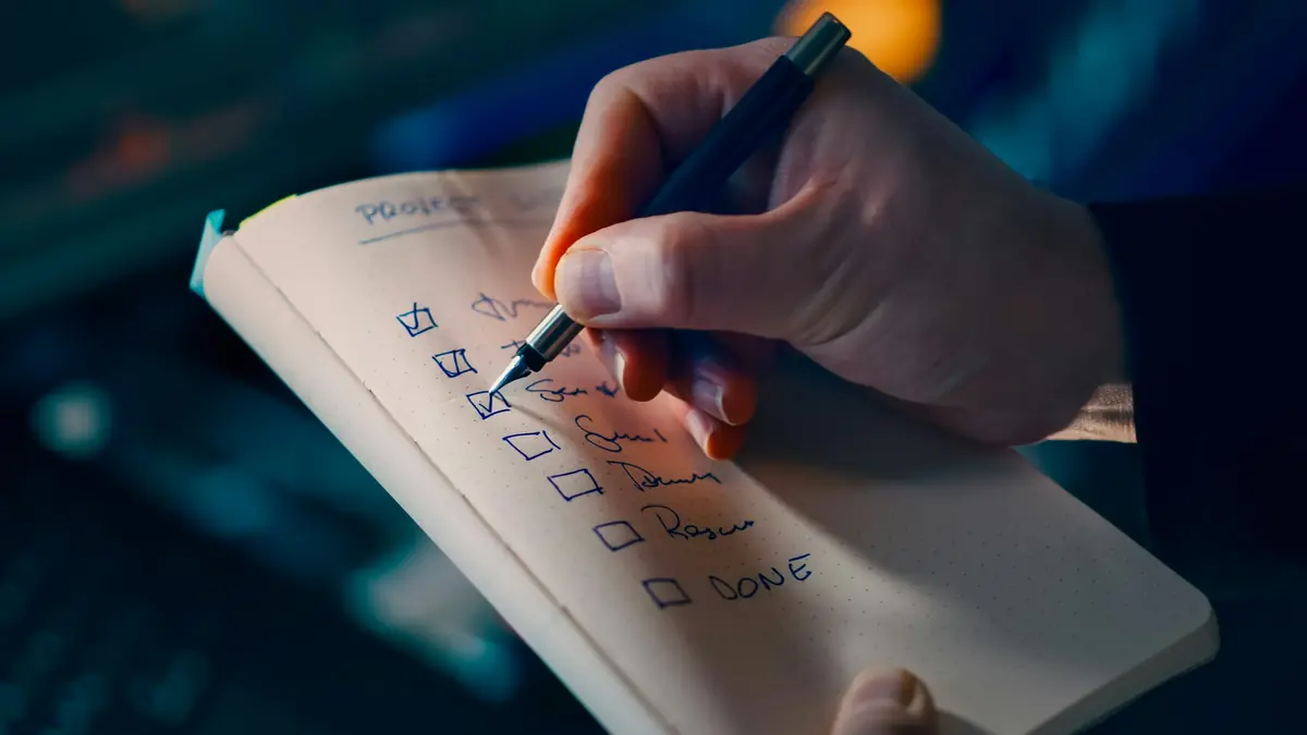 A hand ticking boxes on a handwritten project checklist in a notebook, against a dark blue background