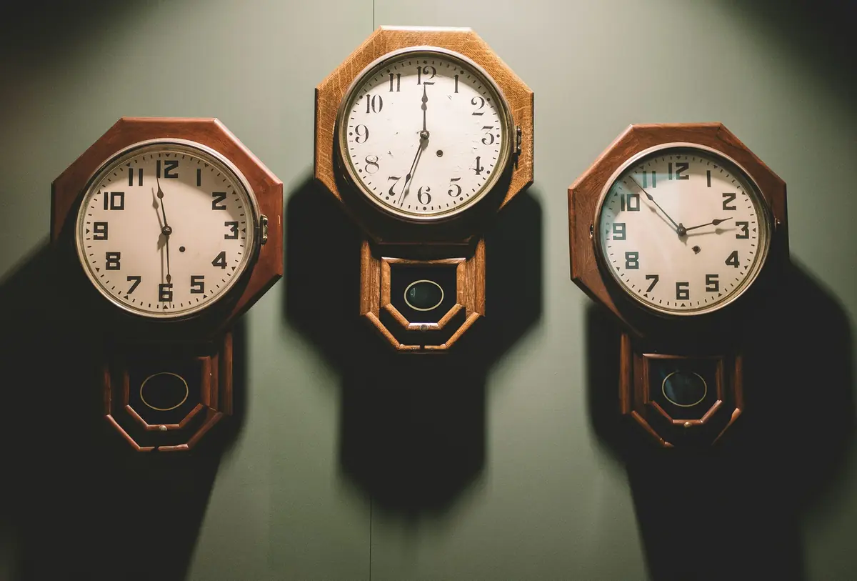 Three wooden pendulum wall clocks showing different times, side by side on a dark green wall