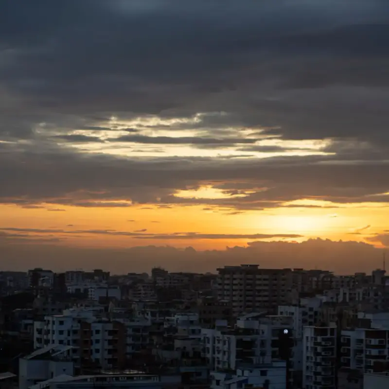 Dhaka rooftops at sunset, dark apartment blocks under heavy grey clouds with a band of orange light on the horizon