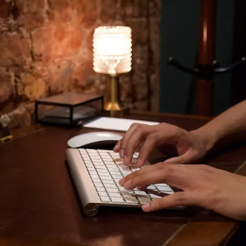 Hands typing on a white keyboard at a dark wooden desk, with a glowing table lamp against a brick wall