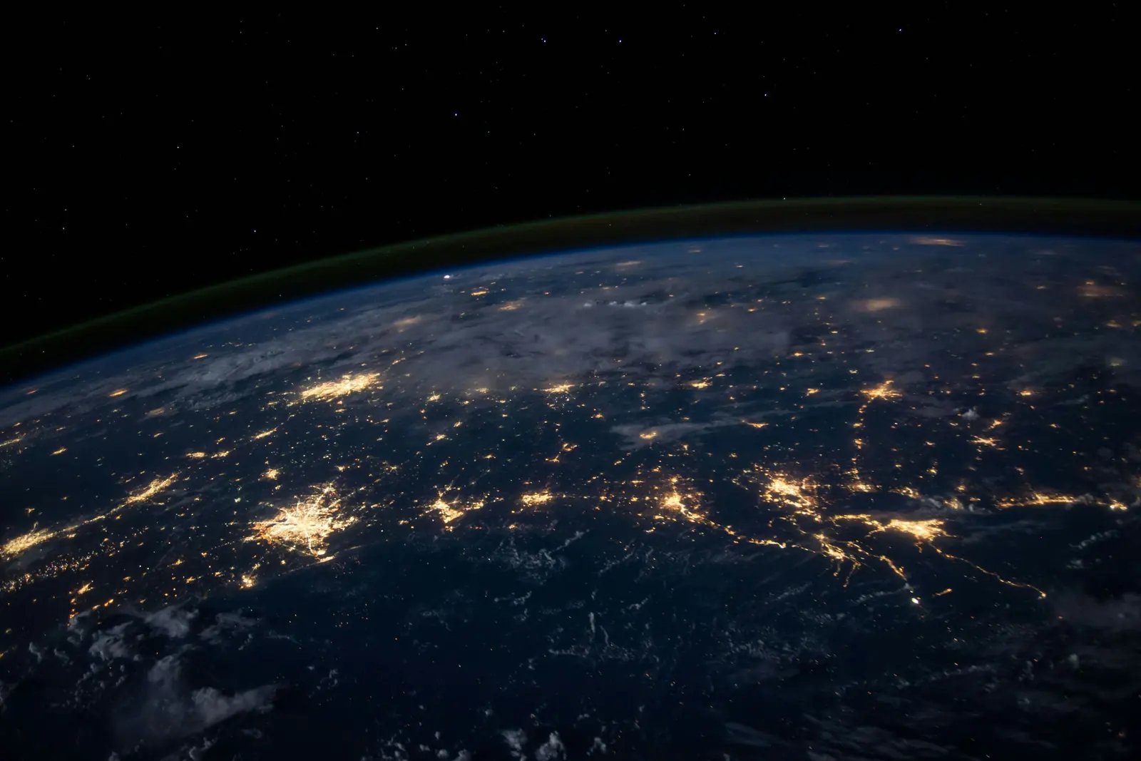 Earth at night from orbit, city lights scattered across the dark surface beneath a thin band of atmosphere and stars