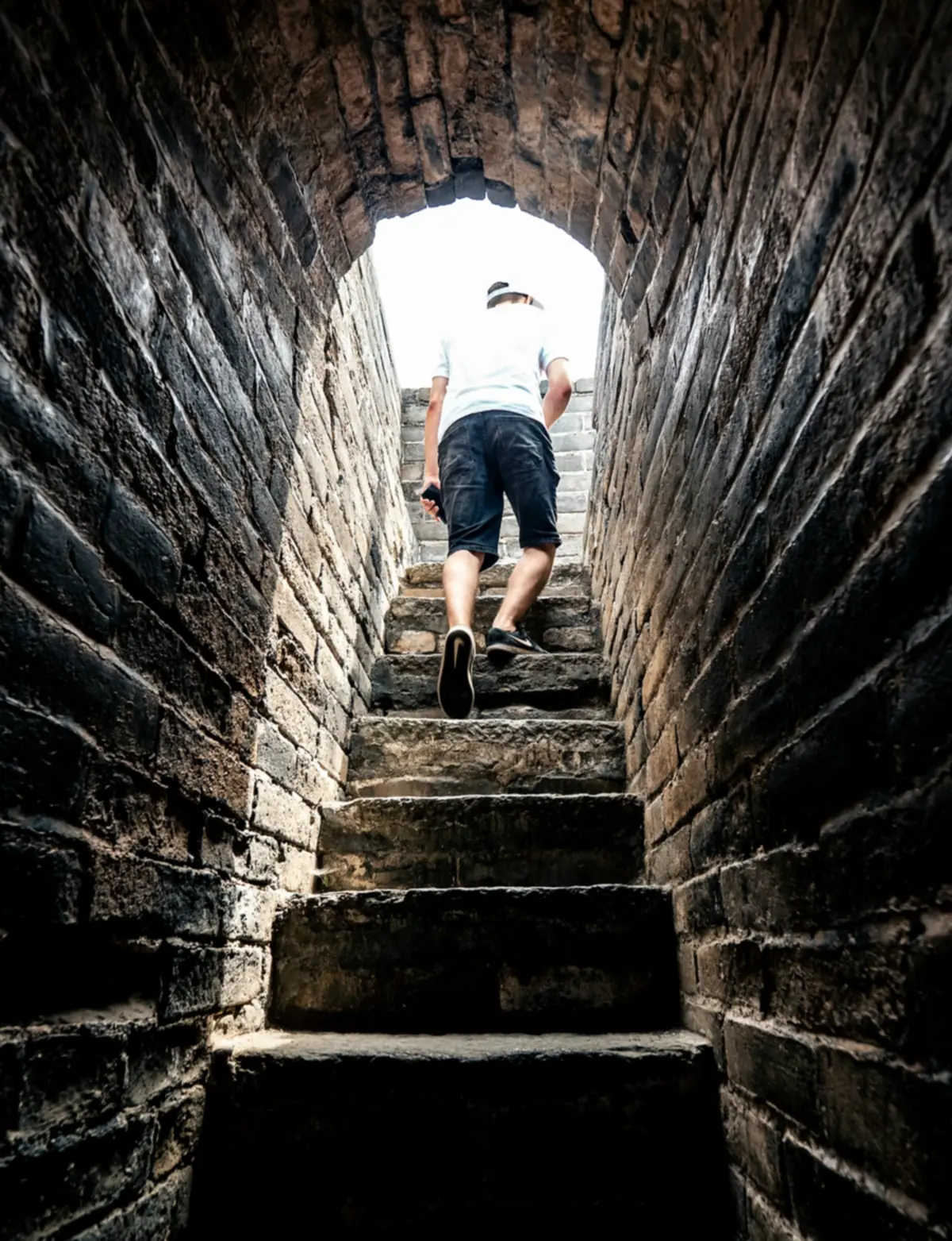A man climbing worn stone steps through a dark brick tunnel towards bright daylight at the top