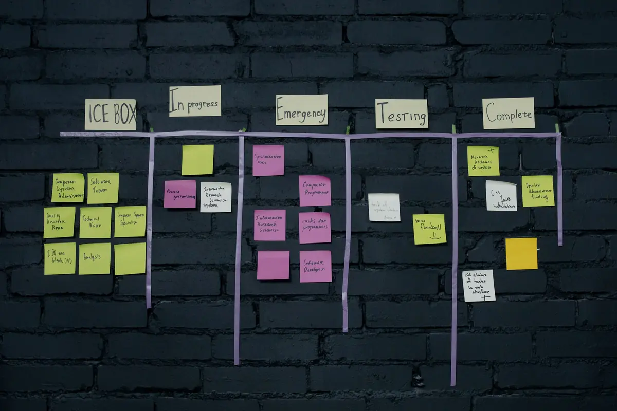 A handmade kanban board on a dark brick wall, with taped lanes and coloured sticky notes under handwritten column headings