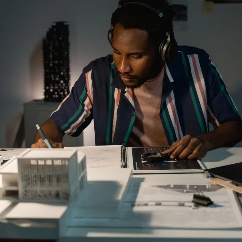 A man in a striped shirt and headphones sketching at a desk with white architectural models, in a dim room at night