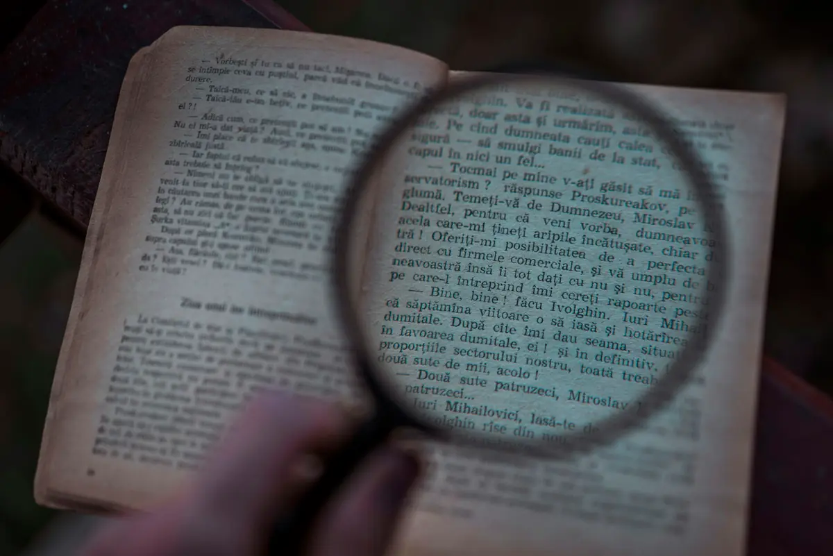 A hand holding a magnifying glass over the yellowed page of an old printed book in dim light