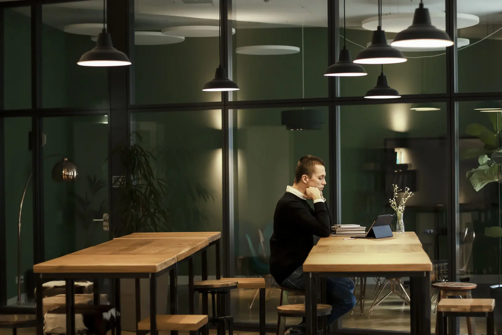 A modern office in the evening with black pendant lamps, glass partitions and oak tables, one man working on a tablet