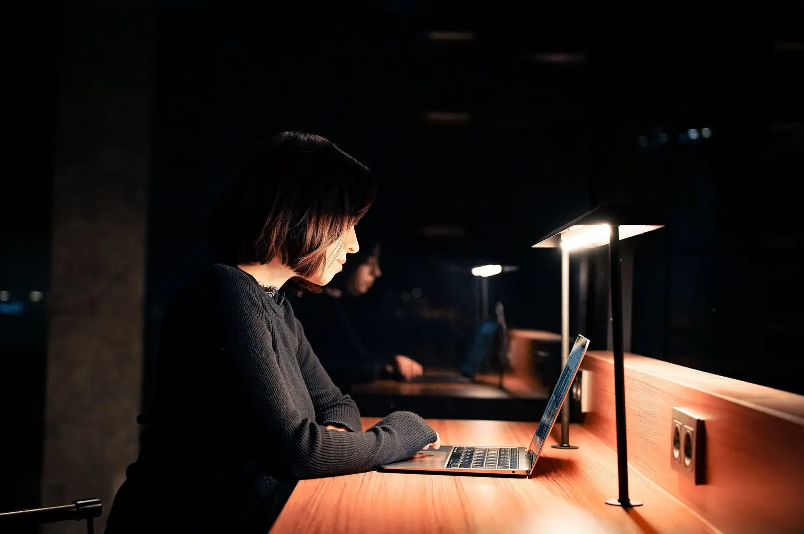 A woman working on a laptop at a wooden counter at night, lit only by a slim desk lamp