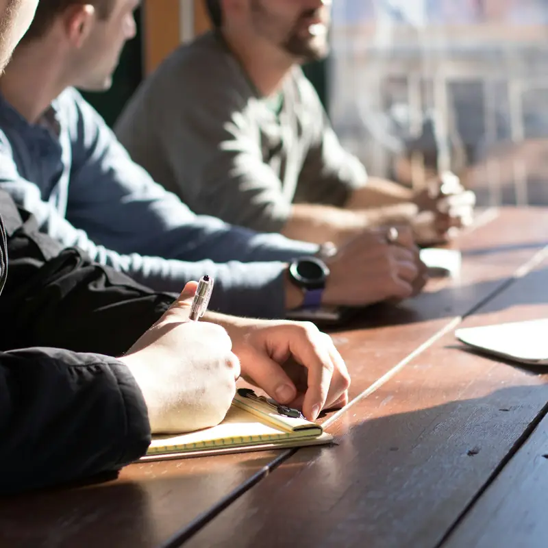 People along a long wooden table in warm window light, taking notes on paper pads during a meeting
