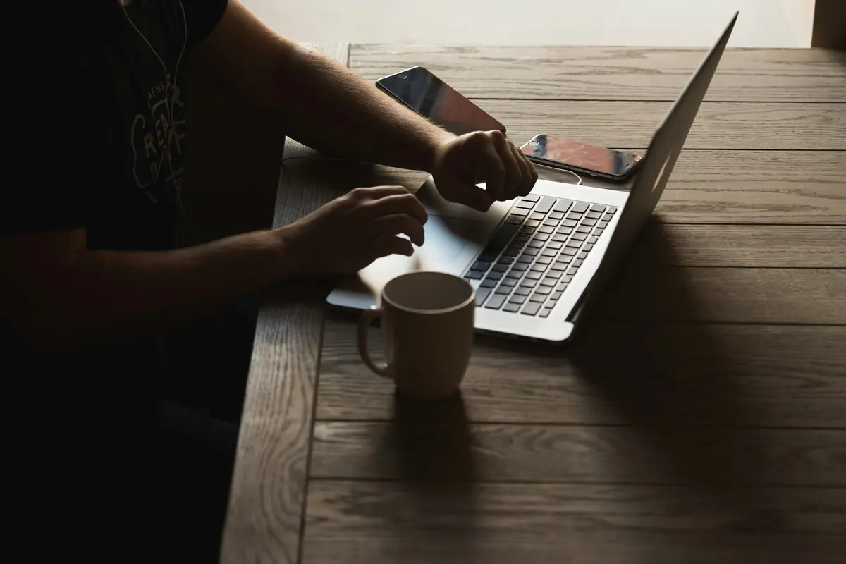 Side view of hands typing on a laptop at a dark wooden table, with a mug and two phones beside it