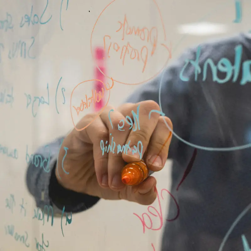 A hand writing on a glass whiteboard with an orange marker through layers of handwritten words and circled notes