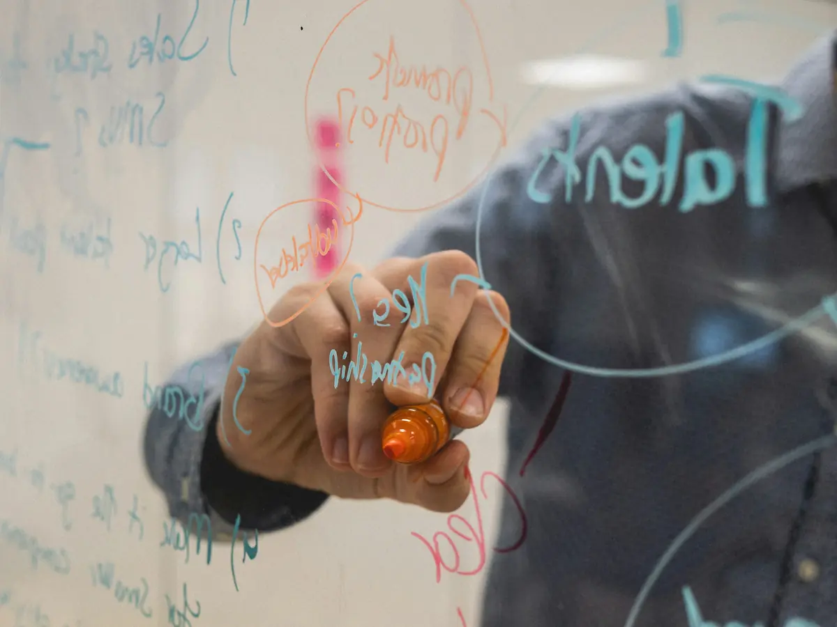 A hand writing on a glass whiteboard with an orange marker through layers of handwritten words and circled notes