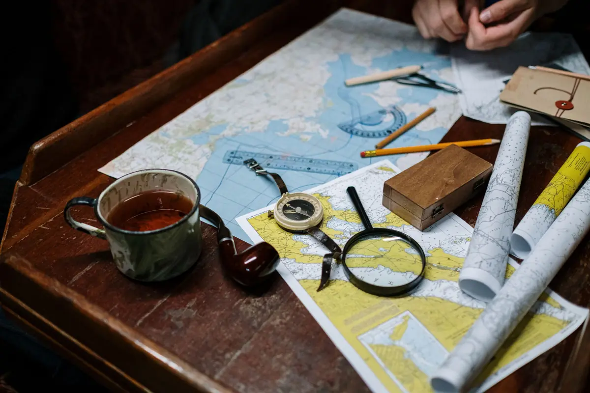 Nautical charts and rolled maps spread over an old wooden desk with a compass, magnifying glass, pencils, a mug of tea and a pipe